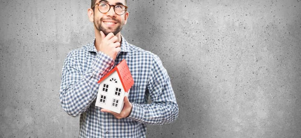 Homme souriant et pensif tenant une maquette de maison face à un mur en béton, illustrant une réflexion sur vos droits et obligations face à un logement encombré ou abandonné.