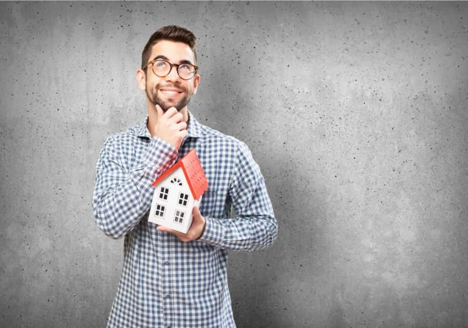 Homme souriant et pensif tenant une maquette de maison face à un mur en béton, illustrant une réflexion sur vos droits et obligations face à un logement encombré ou abandonné.